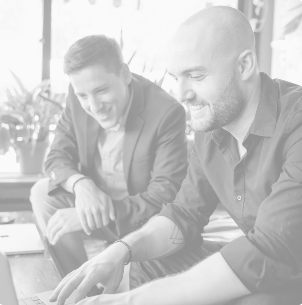 Deux hommes assis à une table; l’homme au premier plan, barbu et vêtu d’une chemise foncée, montre quelque chose devant lui tandis que l’autre sourit.