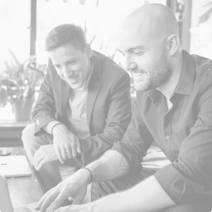 Deux hommes assis à une table; l’homme au premier plan, barbu et vêtu d’une chemise foncée, montre quelque chose devant lui tandis que l’autre sourit.