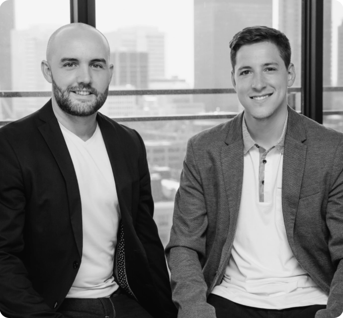 Deux hommes souriants posent côte à côte devant une grande fenêtre donnant sur des immeubles urbains.