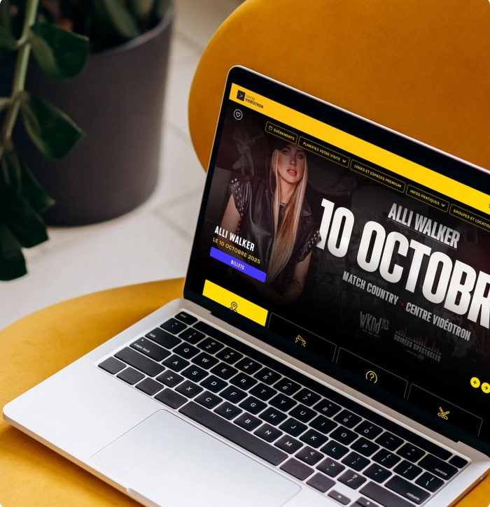 Ordinateur portable affichant une page Web musicale avec « ALL WALKER » et un grand titre « 10 OCTOB… », posé sur une table jaune à côté d’une plante.
