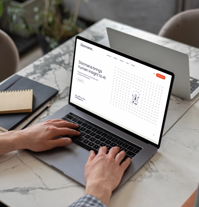 Une personne tape sur un ordinateur portatif affichant une page Web avec un titre comprenant « AI », sur une table en marbre, près d’une tablette, d’un carnet et d’une plante.