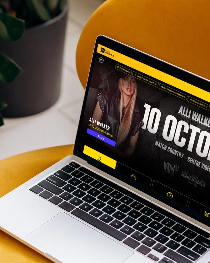 Ordinateur portable affichant une page Web musicale avec « ALL WALKER » et un grand titre « 10 OCTOB… », posé sur une table jaune à côté d’une plante.