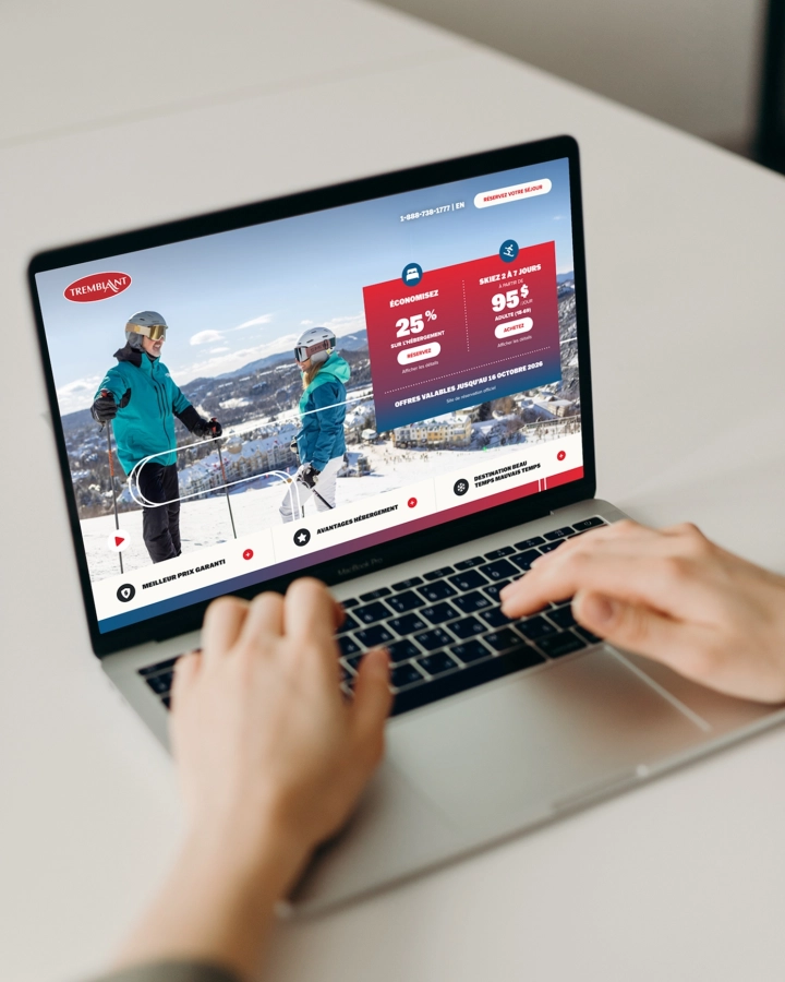 Une personne utilise un ordinateur portable affichant une page Web promotionnelle sur le ski, avec deux skieurs sur une piste enneigée et des offres mises en évidence en rouge.