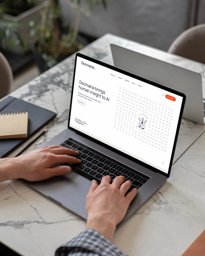 Une personne tape sur un ordinateur portatif affichant une page Web avec un titre comprenant « AI », sur une table en marbre, près d’une tablette, d’un carnet et d’une plante.
